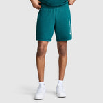 A person wearing Champion 7" Jersey Shorts, Script Logo in teal—crafted from heritage cotton with a white Champion script on the left leg—paired with a matching shirt, white socks, and white sneakers, stands against a plain light gray background.