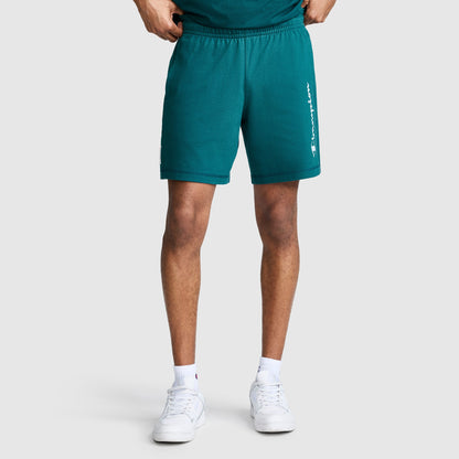 A person wearing Champion 7" Jersey Shorts, Script Logo in teal—crafted from heritage cotton with a white Champion script on the left leg—paired with a matching shirt, white socks, and white sneakers, stands against a plain light gray background.