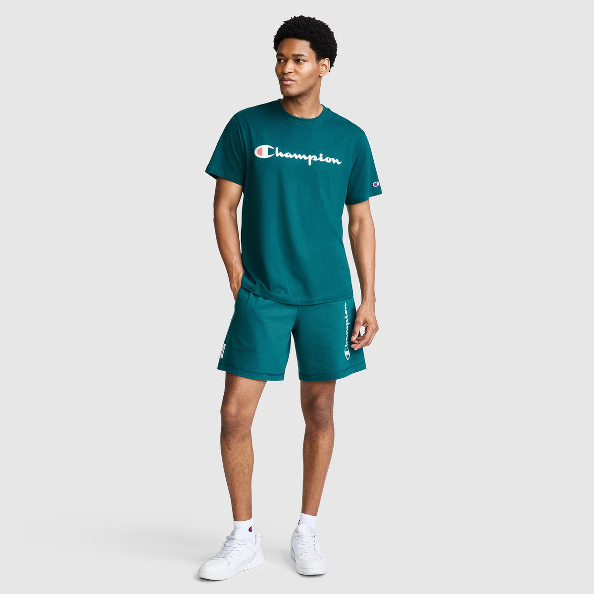 A man stands against a plain light gray background, wearing matching teal Champion 7" Jersey Shorts with the signature script logo and a coordinating t-shirt, both crafted from soft heritage cotton. He completes the look with white sneakers, while the Champion branding subtly accents his outfit.