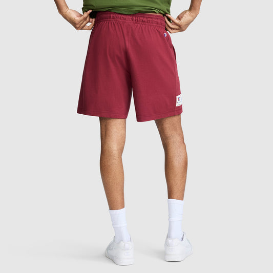 A person stands facing away, wearing a green shirt, white socks, white sneakers, and maroon Champion 7" Jersey Shorts with Script Logo against a light gray background.