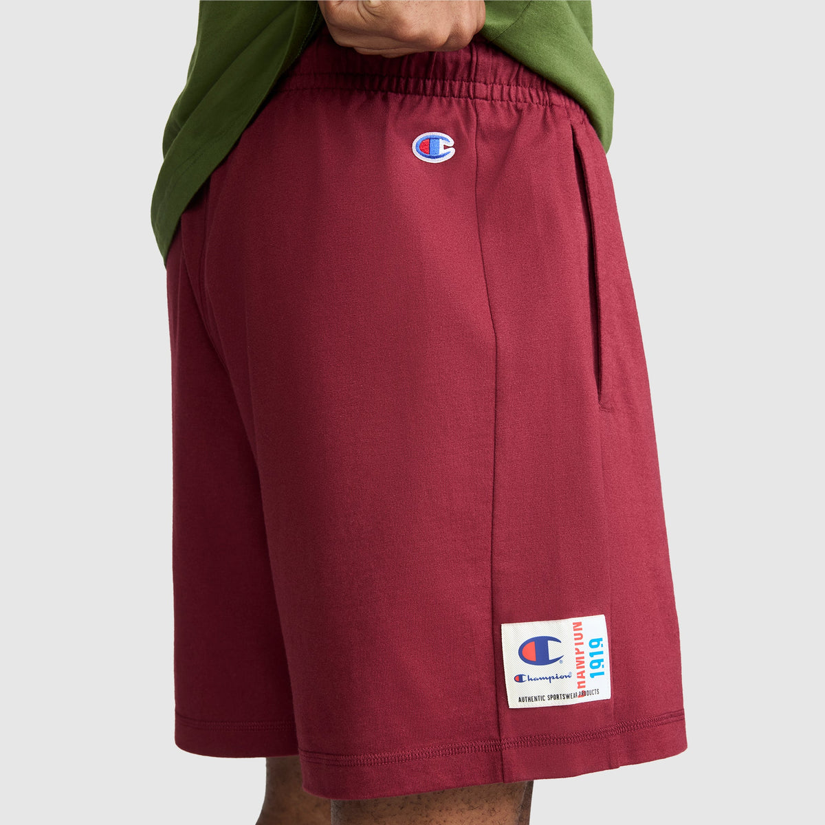 A person wearing maroon Champion 7" Jersey Shorts with the script logo and a green shirt, with one hand in the shorts pocket. The Champion brand logo and a patch with text are visible on the shorts. The background is plain light gray.