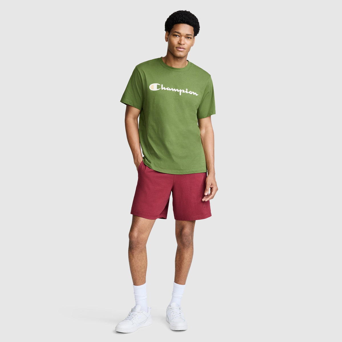 A man stands against a plain light gray background wearing a green Champion t-shirt, Champion 7" Jersey Shorts with Script Logo in red, white socks, and white sneakers.