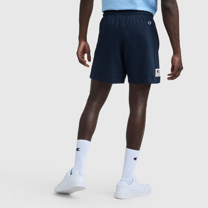 A person faces away, wearing a light blue heritage cotton jersey shirt, Champion 7″ Jersey Shorts, Script Logo in navy, white crew socks with Champion script, and white sneakers against a plain light background.