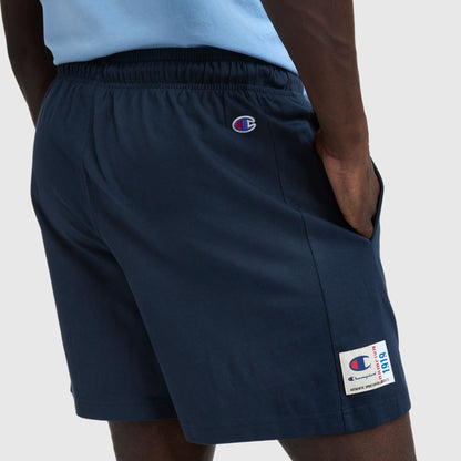 A person wearing navy blue 7″ Jersey Shorts, Script Logo by Champion with hands in pockets. The shorts show a Champion script at the waist and a cotton jersey label patch above the left hem.