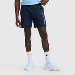 A person wears navy 7″ Jersey Shorts, Script Logo by Champion, white Champion crew socks, and white sneakers against a light gray background. Only the lower half of their body is visible.