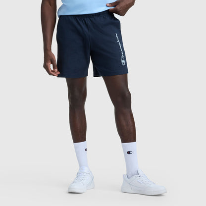 A person wears navy 7″ Jersey Shorts, Script Logo by Champion, white Champion crew socks, and white sneakers against a light gray background. Only the lower half of their body is visible.