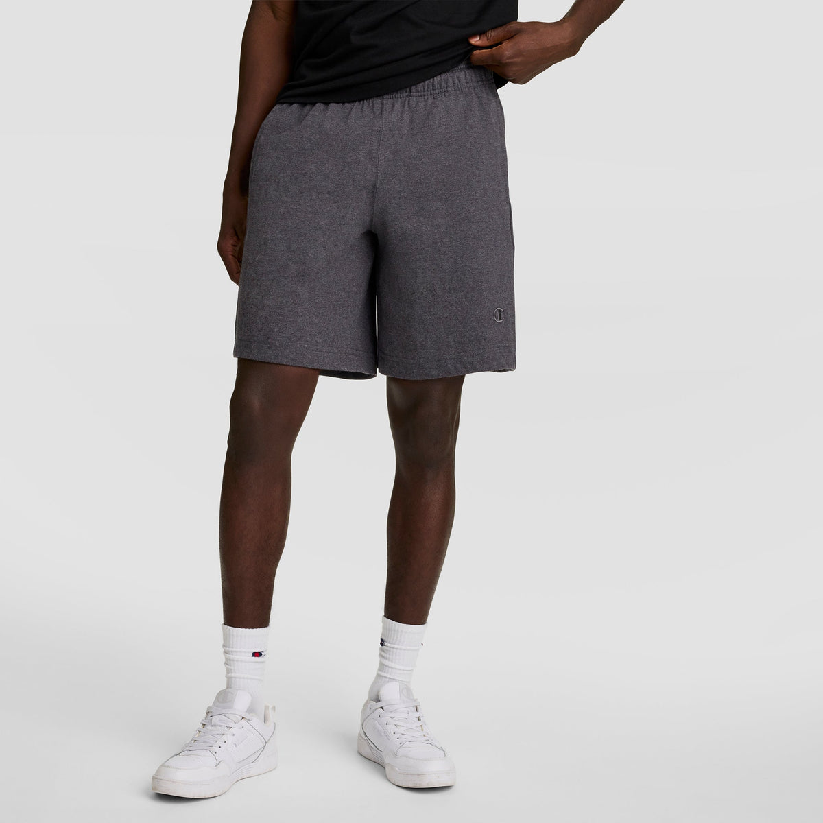 A person in a black t-shirt, Champion 7" Jersey Short with C Logo in gray, white crew socks, and white sneakers stands with one hand in their pocket against a plain light background.