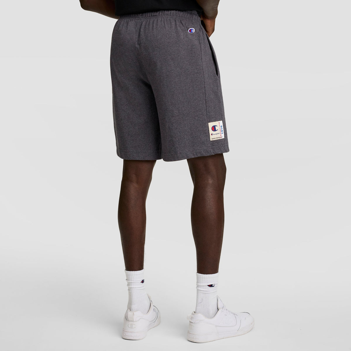 A person stands facing away, wearing Champion 7" Jersey Short with C Logo in dark gray, paired with white crew socks featuring a small logo and white sneakers against a plain light background.