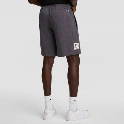 A person stands facing away, wearing Champion 7" Jersey Short with C Logo in dark gray, paired with white crew socks featuring a small logo and white sneakers against a plain light background.