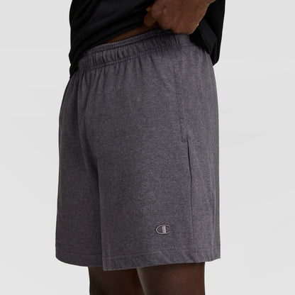 A person wearing Champion 7" Jersey Short, C Logo in dark gray, featuring an elastic waistband and small logo near the hem, stands against a plain light background.