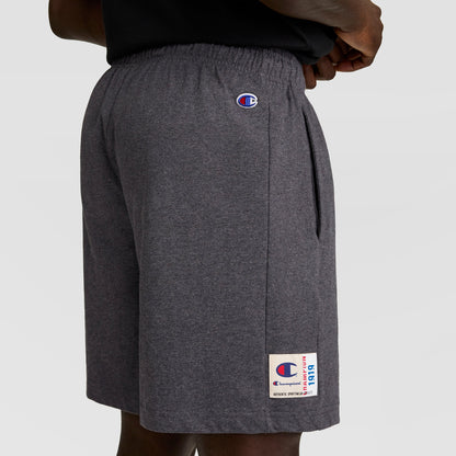 A person wears dark gray Champion 7" Jersey Shorts, featuring the C logo on the left thigh and a small patch near the hem, with hands on hips against a plain light gray background.
