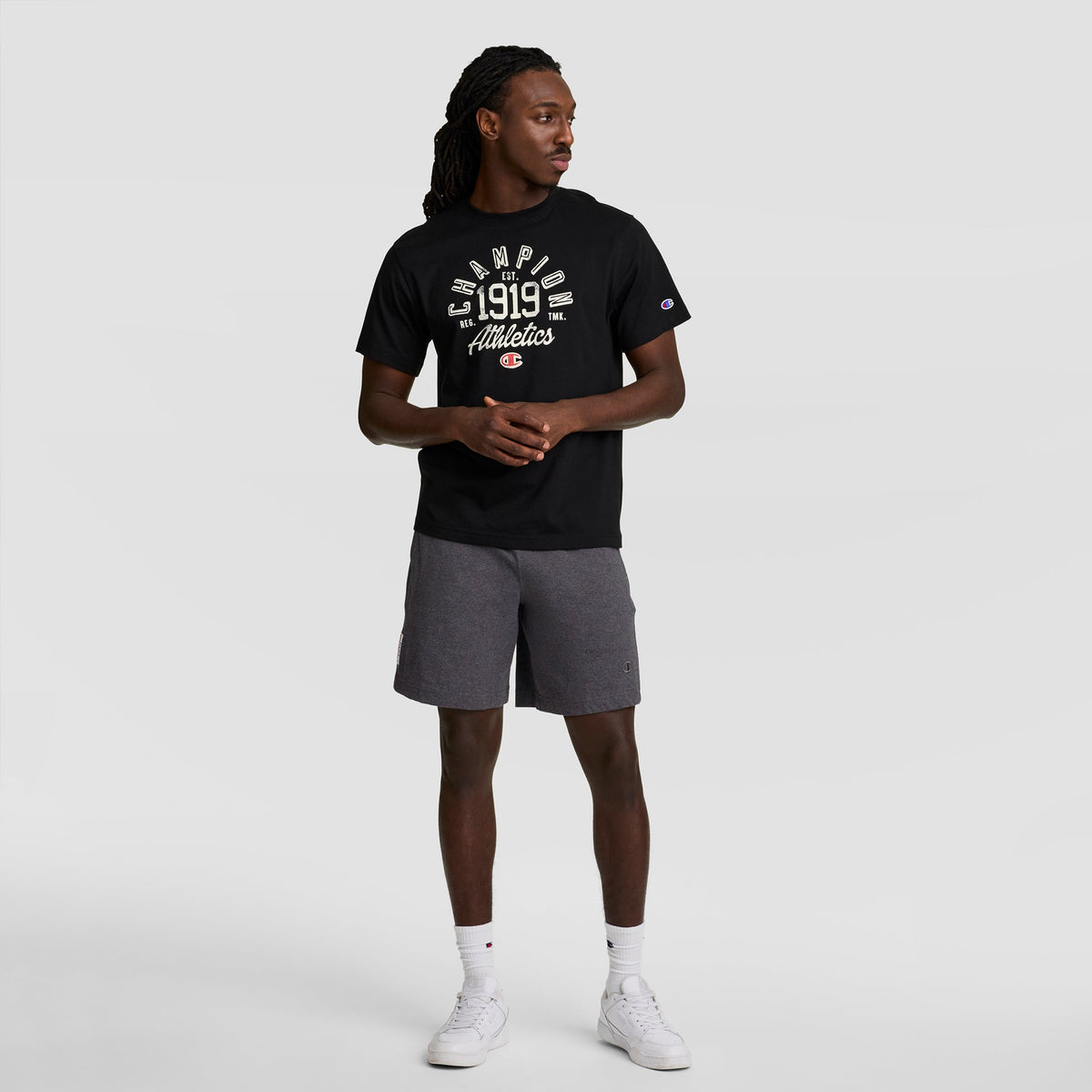 A man stands against a plain background wearing a black Champion 1919 Athletics t-shirt and Champion 7" Jersey Short with C Logo in gray, paired with white sneakers and socks. He looks left with his hands clasped in front of him.