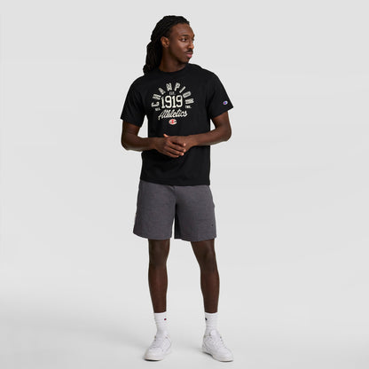 A man stands against a plain background wearing a black Champion 1919 Athletics t-shirt and Champion 7" Jersey Short with C Logo in gray, paired with white sneakers and socks. He looks left with his hands clasped in front of him.