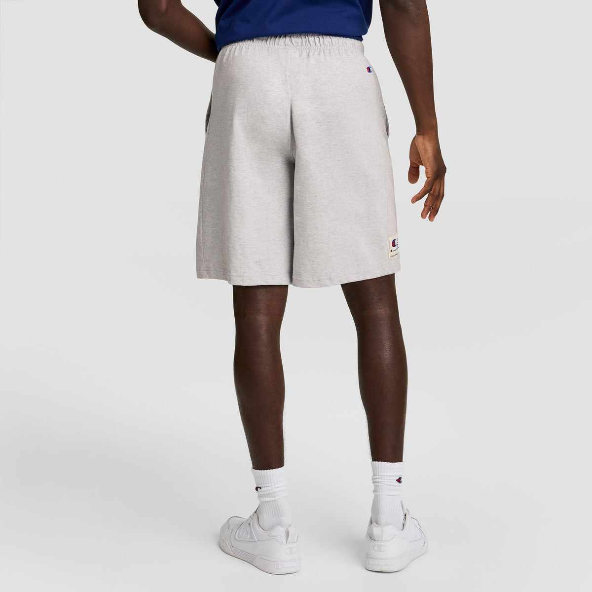 A person stands facing away, wearing Champion 7" Jersey Short with C Logo in light gray, paired with a blue top, white socks, and white sneakers against a plain light background.