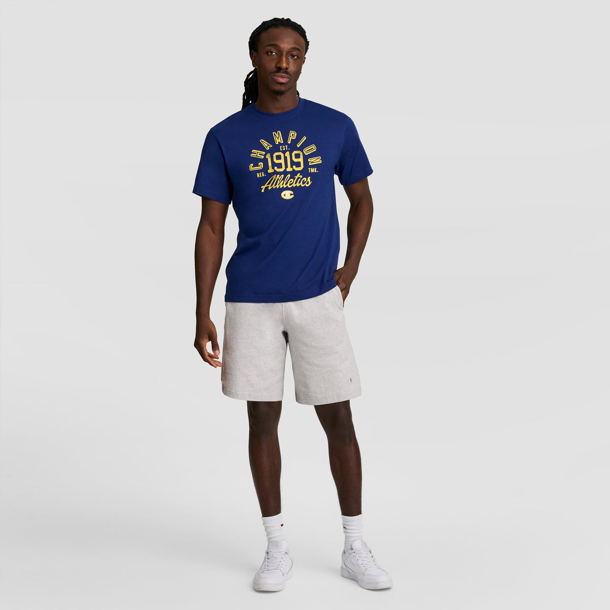 A man faces forward in a blue Champion 1919 Athletics T-shirt, light gray Champion 7" Jersey Shorts with C Logo, and white sneakers against a plain light gray background.