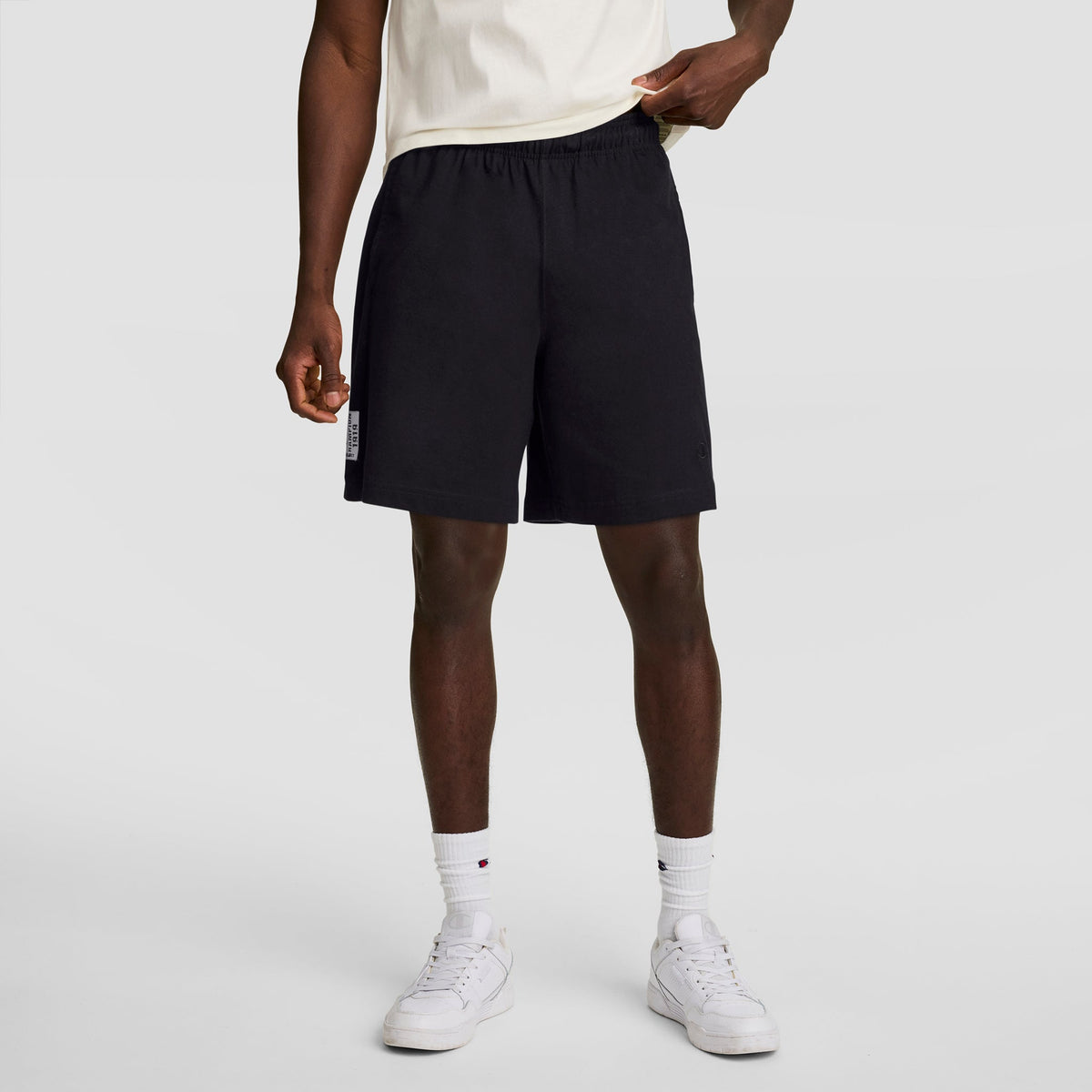 A person in a white cotton jersey t-shirt, Champion 7" Jersey Shorts with C Logo in black, white crew socks, and white sneakers stands against a plain gray background. Their face is not visible.