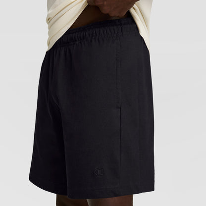 A person in a light shirt lifts it to reveal Champion's 7" Jersey Short, C Logo—black men’s shorts made from soft cotton jersey with an elastic waistband and a small embroidered logo by the hem for classic Champion style.