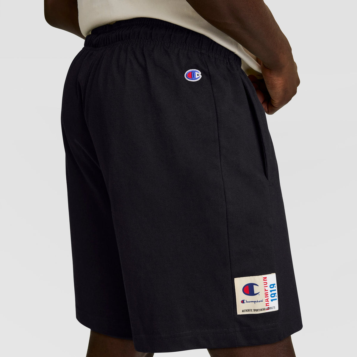 A person wears Champion 7" Jersey Short, C Logo—black men’s shorts with the Champion logo on the left thigh and a white cotton jersey label near the hem. Their right hand rests on their hip.
