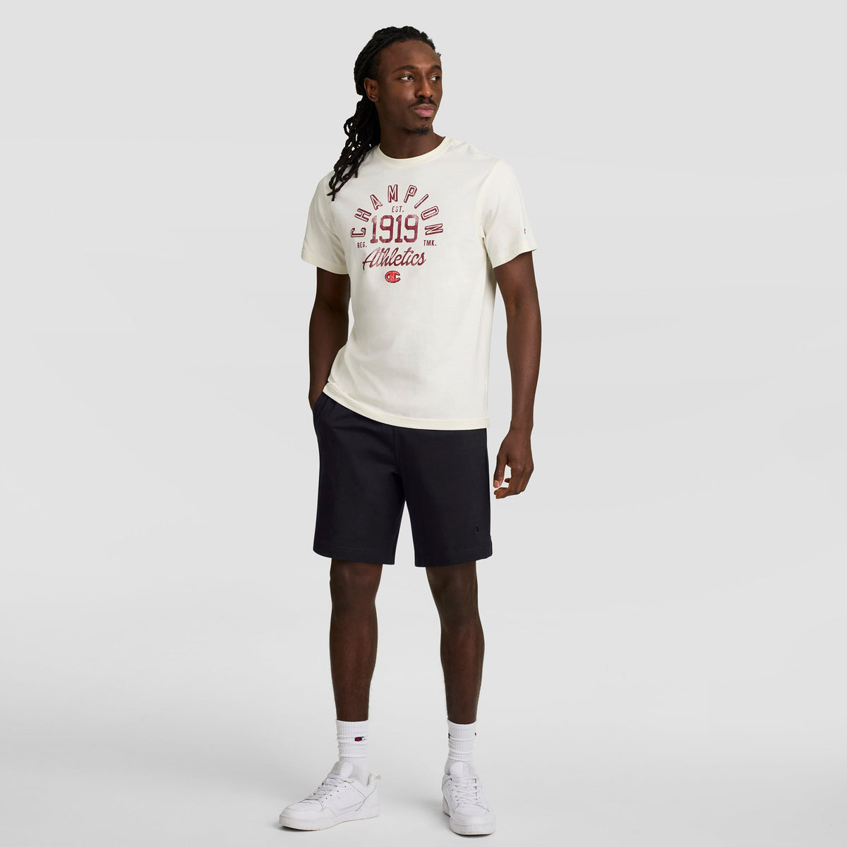 A man with long hair stands against a plain background in a white cotton Champion Athletics 1919 T-shirt, Champion 7" Jersey Short with C Logo (black), white socks, and white sneakers, one hand in his pocket.