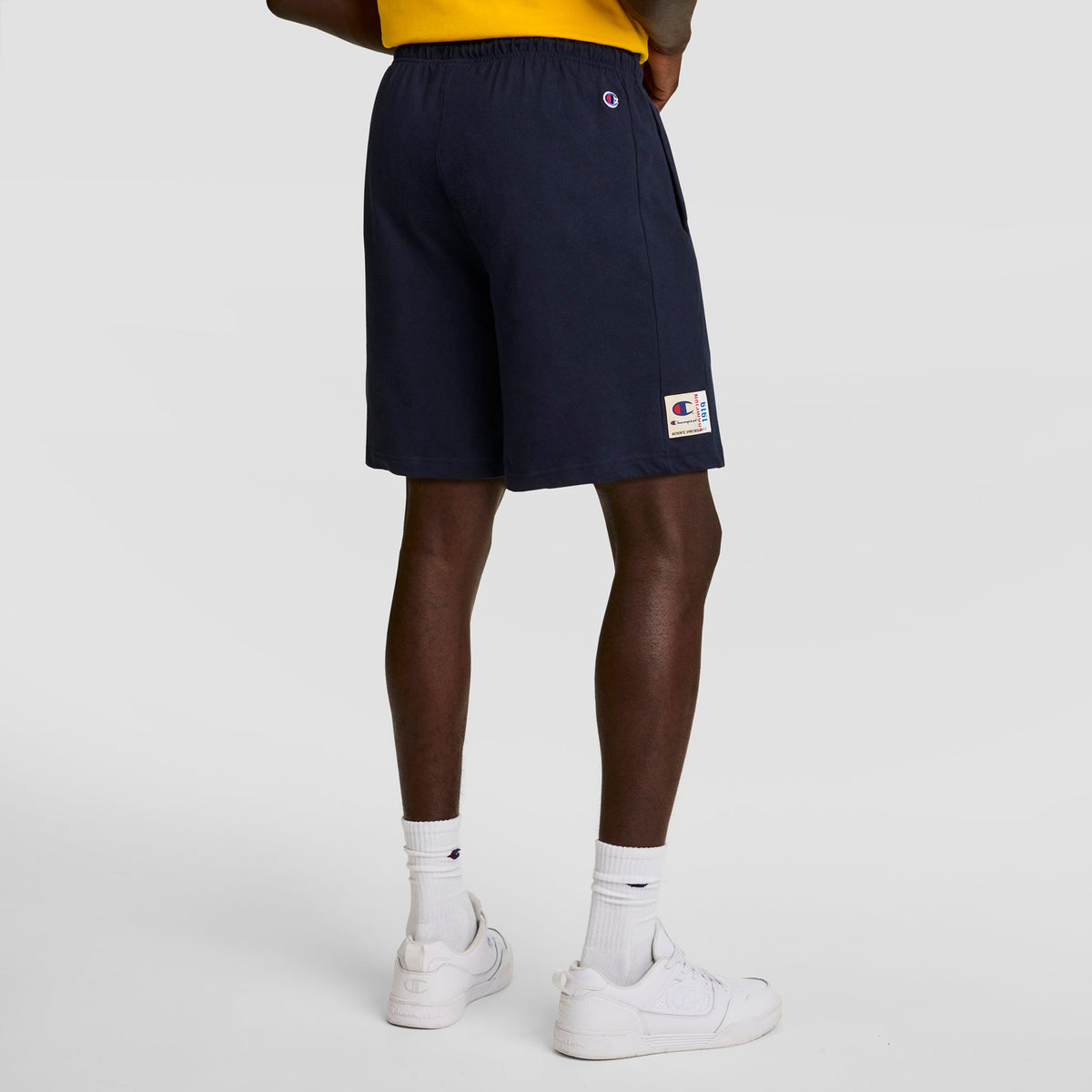 Someone is seen from behind in a yellow shirt, white socks, and white sneakers, wearing Champion 7" Jersey Short with C Logo, standing against a plain light background.