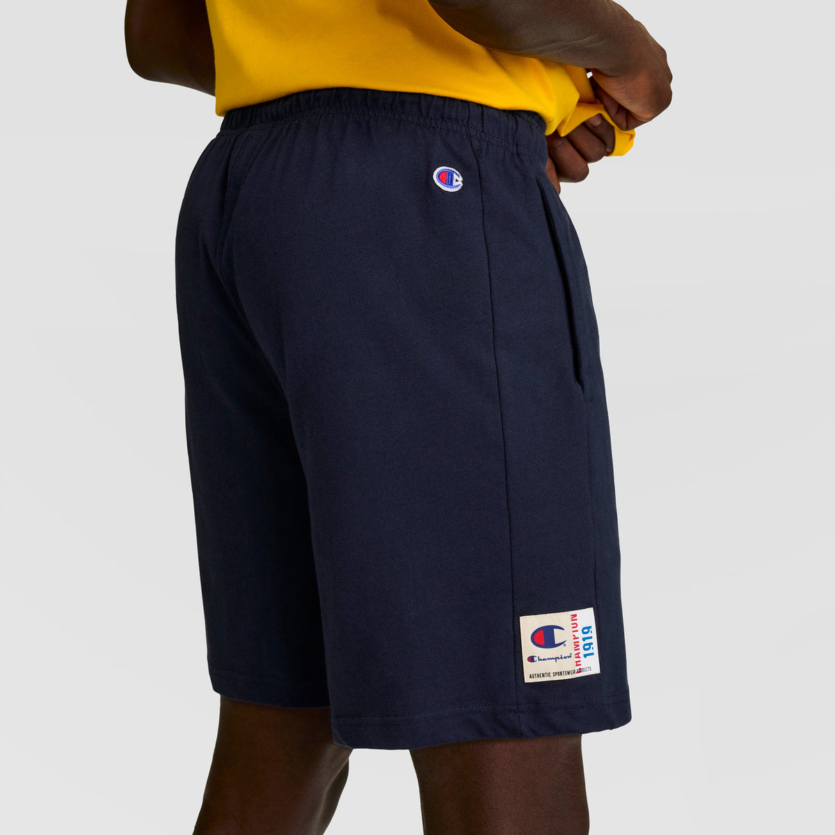 A person poses sideways with one hand on their hip, wearing Champion’s 7" Jersey Short with a visible C Logo in navy blue, paired with a yellow shirt. The men’s athletic shorts by Champion catch the eye.