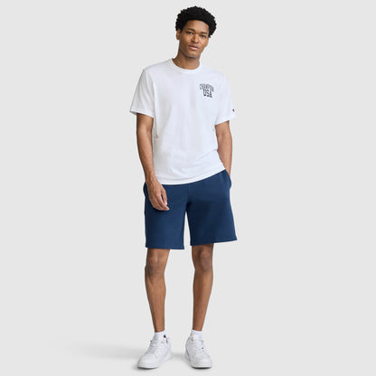 A person stands facing forward, wearing the Champion Short Sleeve USA T-Shirt—a classic white athletic tee featuring "USA" across the chest—paired with navy blue shorts and white sneakers. The background is plain and light gray.