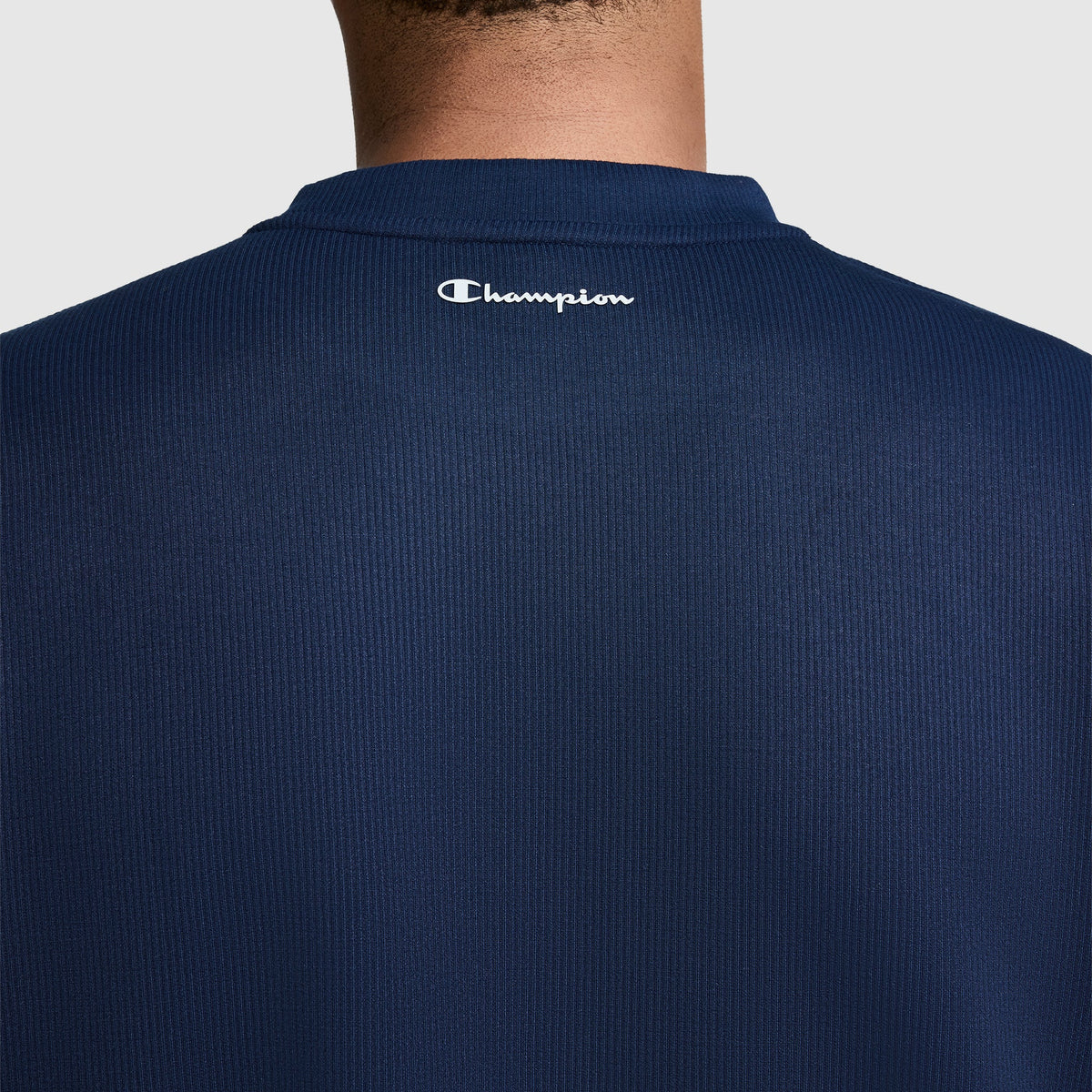 A back view of a person wearing the Champion Sport Long Sleeve Crewneck, C Logo in navy blue ribbed fabric, featuring the white Champion "C" logo printed near the collar. The athletic crewneck provides a comfortable fit, with the wearer's neck and upper back visible.