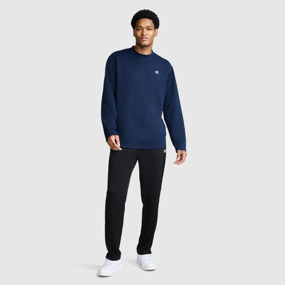 A man stands against a plain background wearing a navy blue Champion Sport Long Sleeve Crewneck, C Logo top, black pants, and white sneakers.