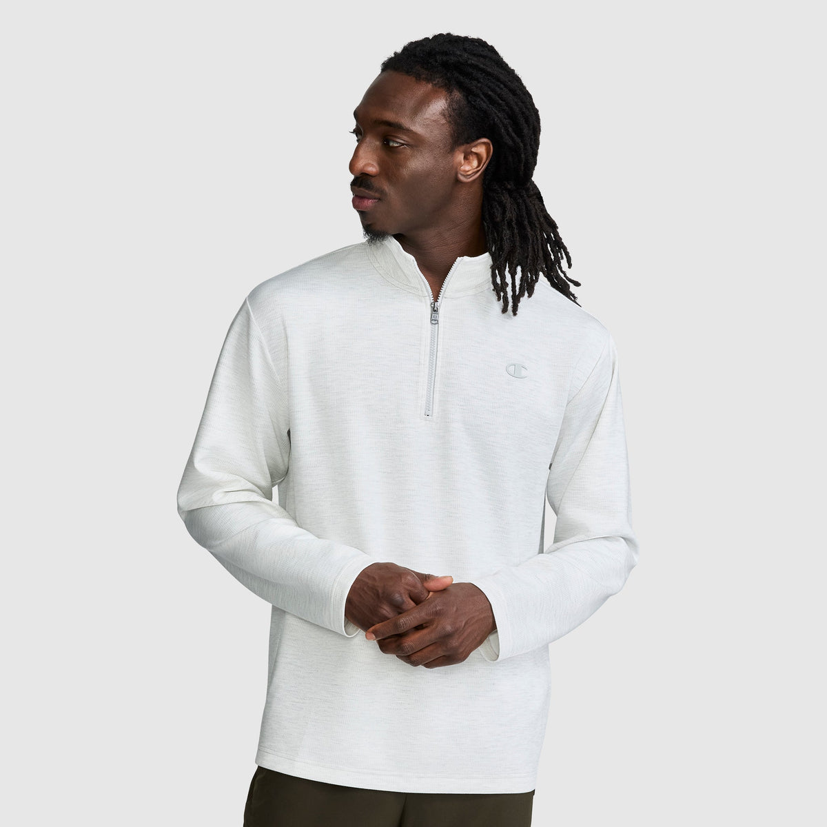 A man with long dreadlocks wears a light gray Champion Sport Long Sleeve 1/4 Zip Mockneck with the C Logo and dark pants, standing against a plain light background while looking to his left.