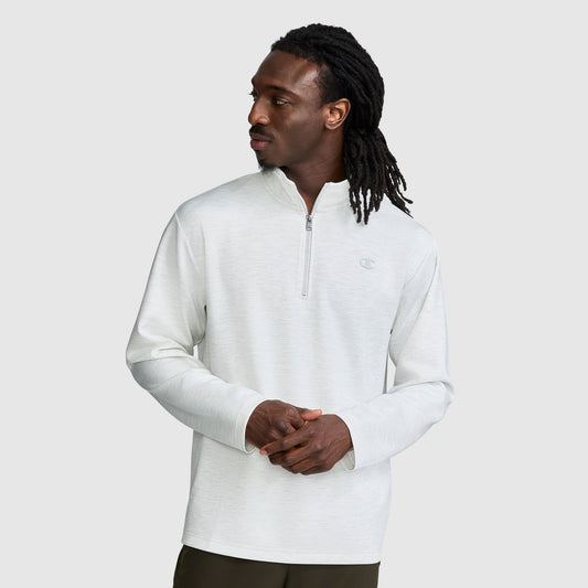 A man with long dreadlocks wears a light gray Champion Sport Long Sleeve 1/4 Zip Mockneck with the C Logo and dark pants, standing against a plain light background while looking to his left.