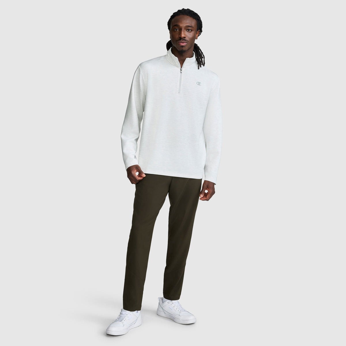 A man stands against a plain background wearing a light gray Champion Sport Long Sleeve 1/4 Zip Mockneck with the C Logo, paired with dark pants and white sneakers. His long hair is styled in twists, and he looks at the camera with a neutral expression.