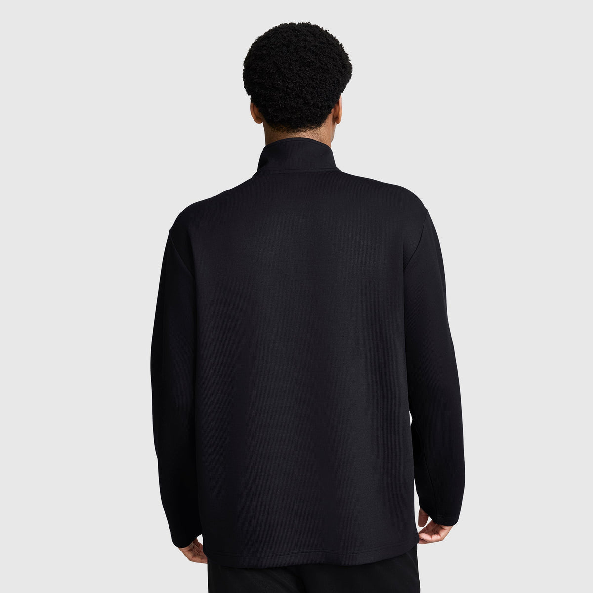 A person with short curly hair is seen from behind, wearing a Champion Sport Long Sleeve 1/4 Zip Mockneck with the C Logo and black pants, standing against a plain light gray background.