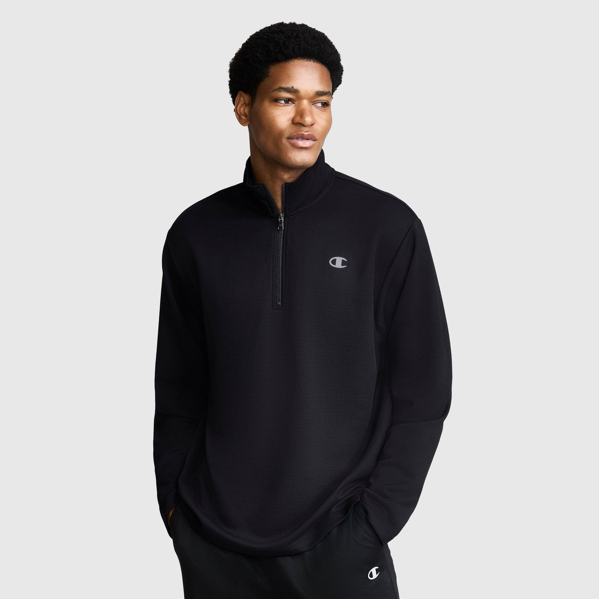 A man wearing a black Champion Sport Long Sleeve 1/4 Zip Mockneck with the C Logo stands with his hands in his pockets, looking slightly to the side against a plain light gray background.