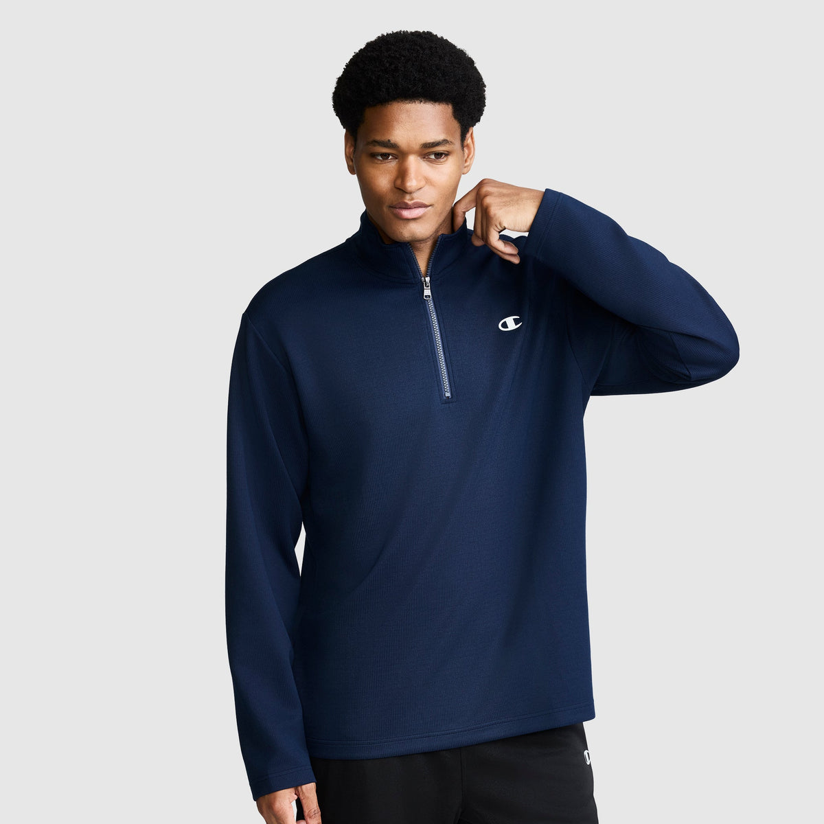 A person wearing a navy blue Champion Sport Long Sleeve 1/4 Zip Mockneck with a C Logo stands against a plain light gray background, touching the zipper with one hand.