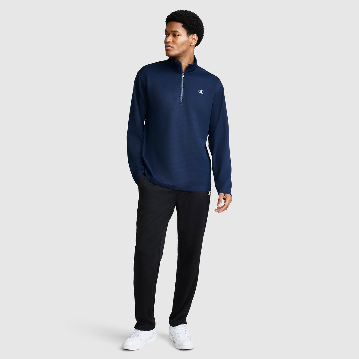 A man stands against a plain light gray background wearing the Champion Sport Long Sleeve 1/4 Zip Mockneck with C Logo, paired with black pants and white sneakers.