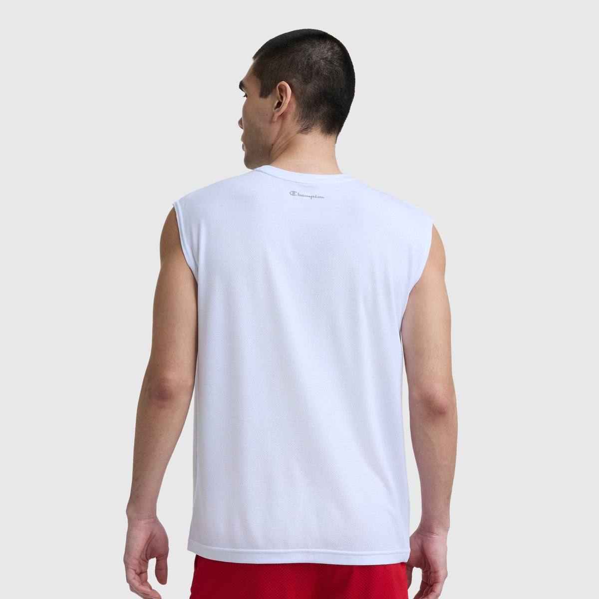 A man with short dark hair stands with his back to the camera, wearing a white Champion Sport Crewneck Muscle Tee, Script Logo, and red shorts against a plain light gray background.