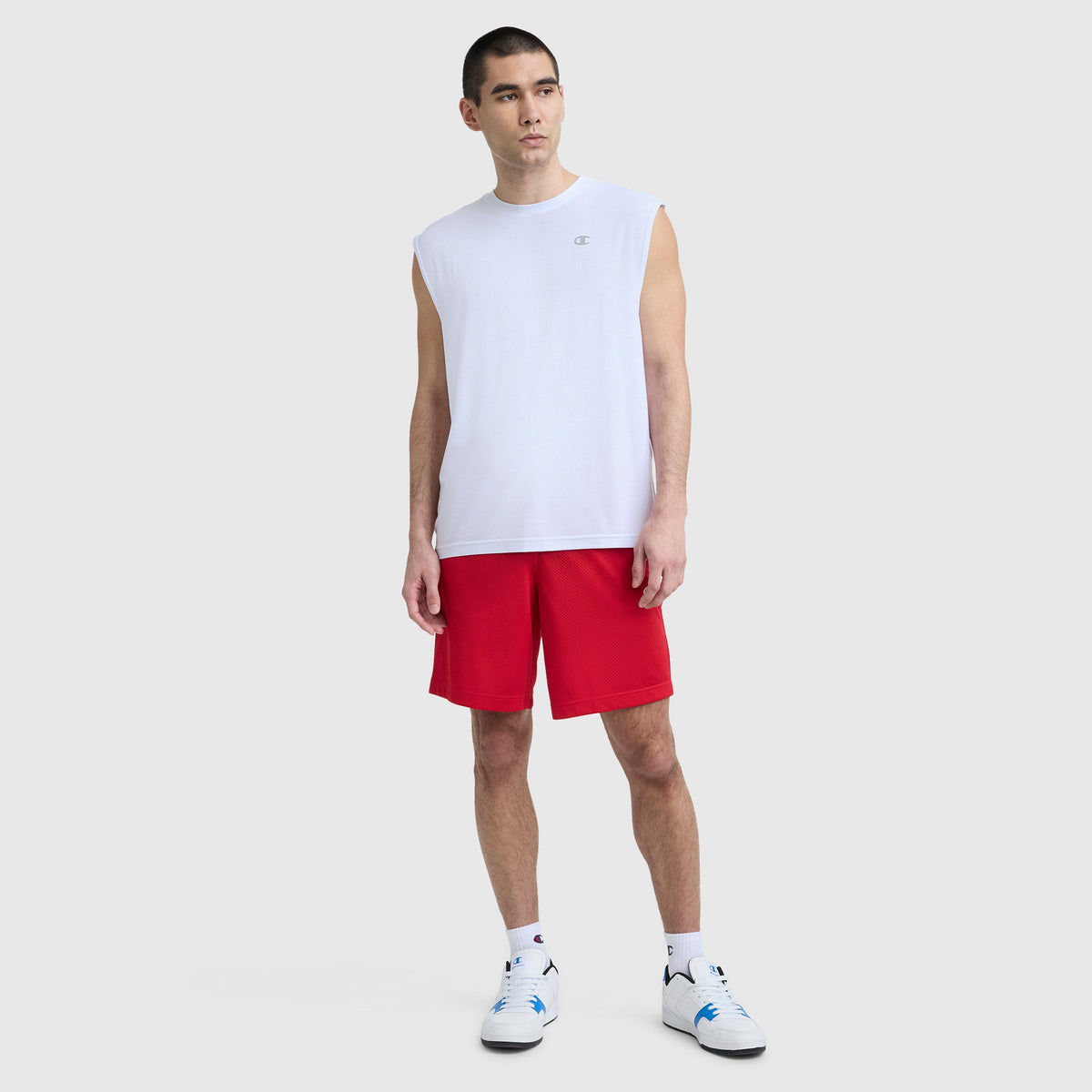 A man stands against a plain background wearing a Champion Sport Crewneck Muscle Tee, Script Logo, red athletic shorts, white socks, and white sneakers with blue accents.
