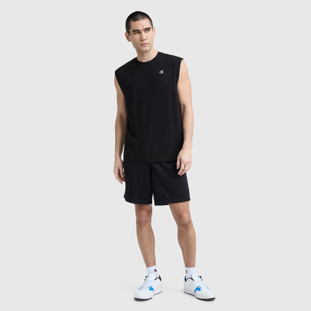 A man stands against a light gray background wearing a Champion Sport Crewneck Muscle Tee with Script Logo, black shorts, white socks, and white sneakers, looking to the side in a relaxed pose.