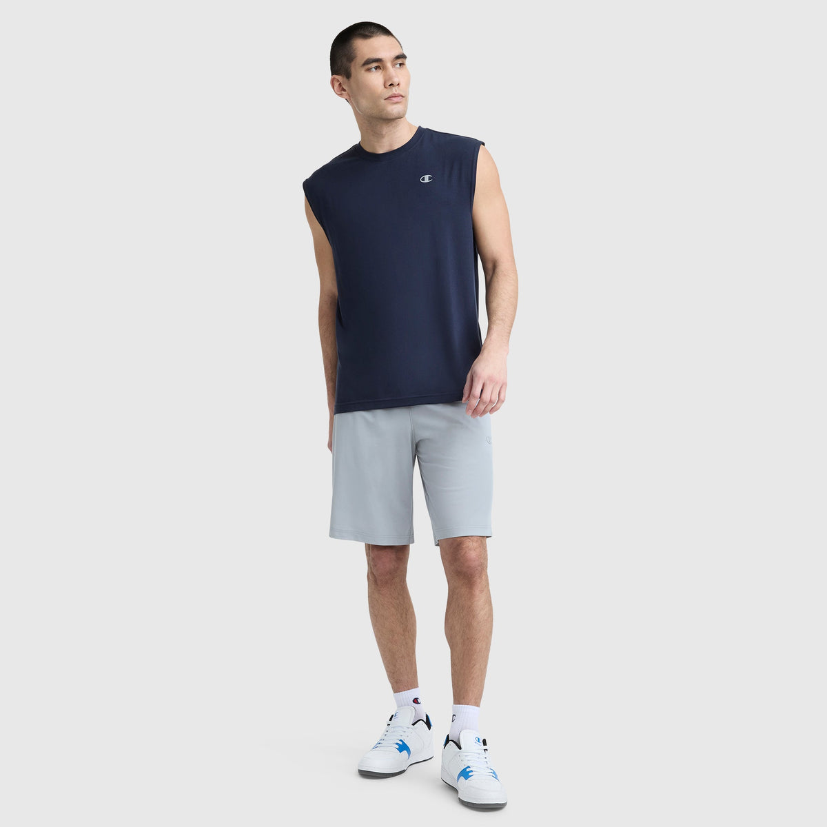 A man wears a Champion Sport Crewneck Muscle Tee, Script Logo, with light gray shorts and white sneakers, standing against a plain light background and looking to the side with one hand in his pocket.