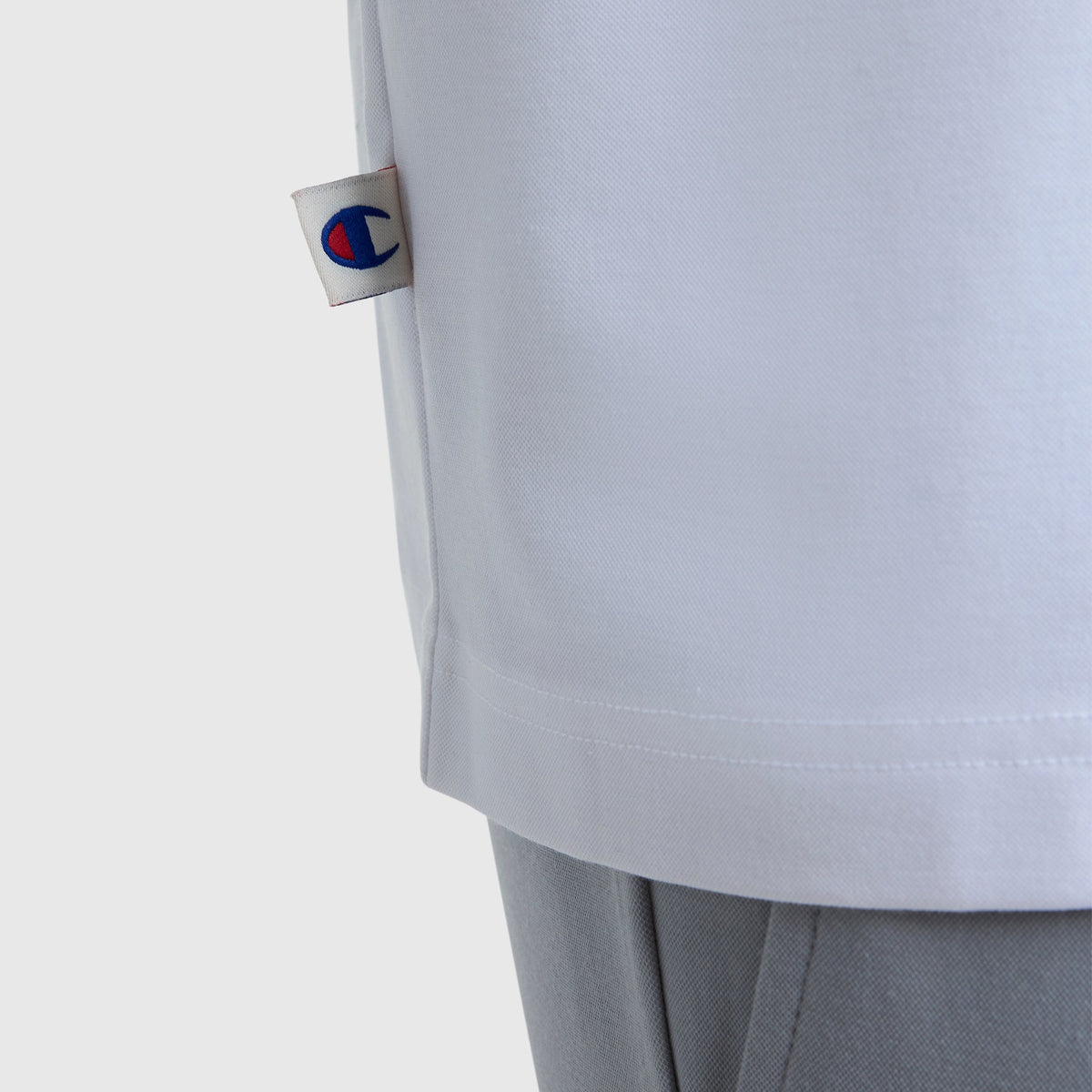 Detail of the Champion Sport Polo, C Logo in white, featuring a small logo tag on the side and styled with light gray pants for an athletic look against a plain white background.