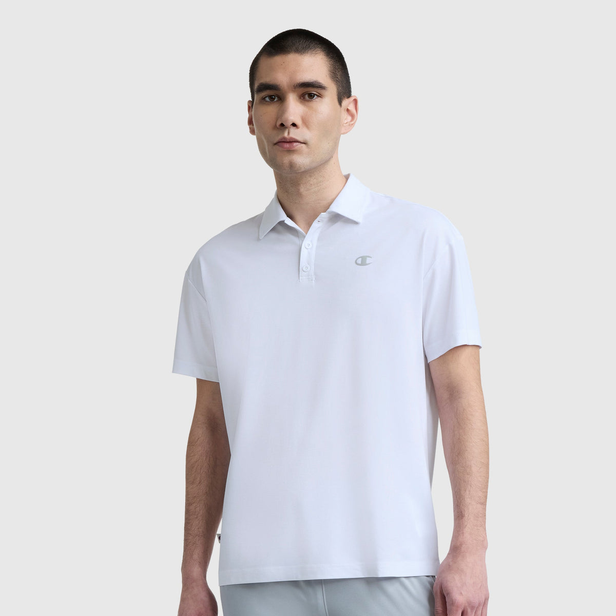 A man with short dark hair wears a plain white Champion Sport Polo, C Logo, featuring a small chest logo for an athletic look as he stands against a light gray background.