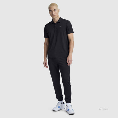 A person with short blond hair wears the Champion Sport Polo, C Logo in black, paired with black joggers and white sneakers with blue accents, standing against a plain light gray background.