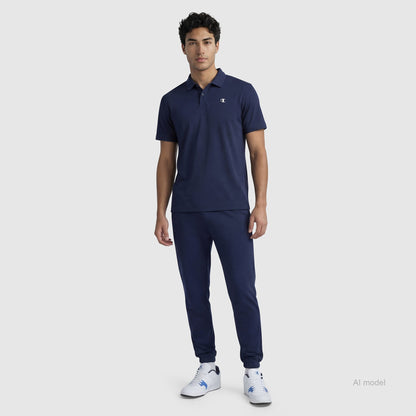 A man stands against a plain background wearing a navy blue Champion Sport Polo, C Logo, matching navy jogger pants, and white sneakers.