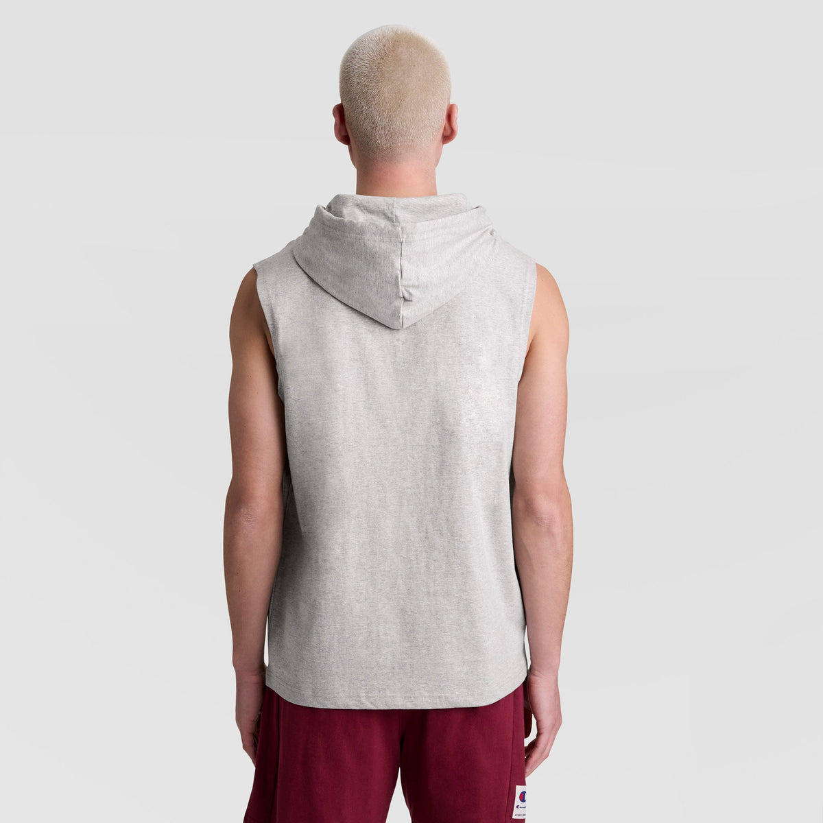A person with short, light blonde hair stands with their back to the camera, wearing a Champion Sleeveless Hoodie with an Embroidered C Logo and burgundy shorts against a plain light gray background.