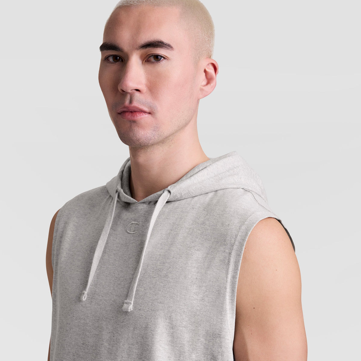 A person with short, light blond hair wears a Champion Sleeveless Hoodie with an Embroidered C Logo and looks toward the camera against a plain light gray background.