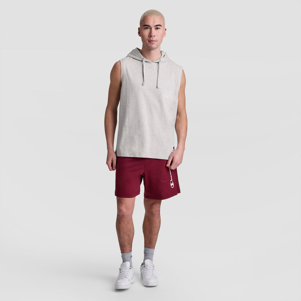 A man with short blond hair wears a Champion Sleeveless Hoodie with an embroidered C logo, maroon shorts featuring a vertical white logo, gray socks, and white sneakers, standing against a plain light gray background.