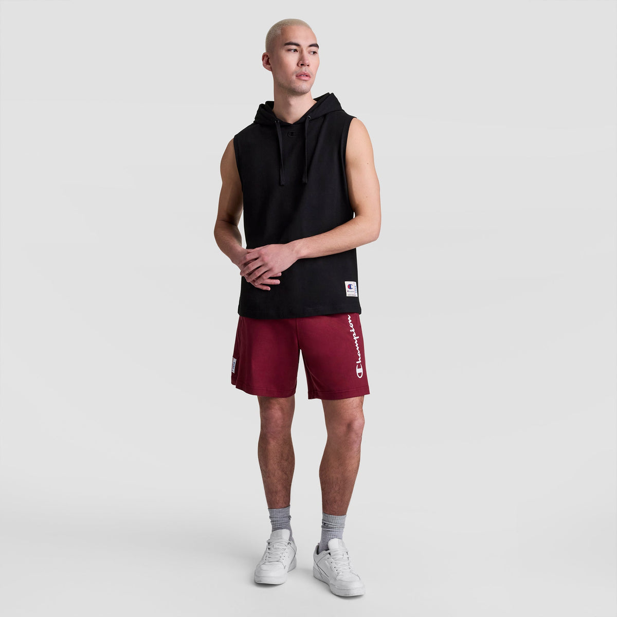 A person in a Champion Sleeveless Hoodie (C Logo), red Champion shorts, gray socks, and white sneakers stands against a plain light gray background.