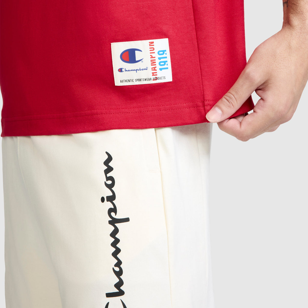 Close-up of a person wearing a red Champion Jersey Sleeveless Hoodie, featuring the C logo, made from soft ring-spun cotton with a rectangular logo tag. They are also wearing white pants with the word "Champion" written in black script along the side and are holding the hem of the hoodie.