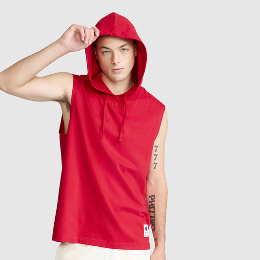 A person wearing the Champion Jersey Sleeveless Hoodie with C Logo in red, crafted from soft ring-spun cotton, and white shorts lifts the hood with one hand. Tattoos decorate their left arm as they stand against a plain light gray background.