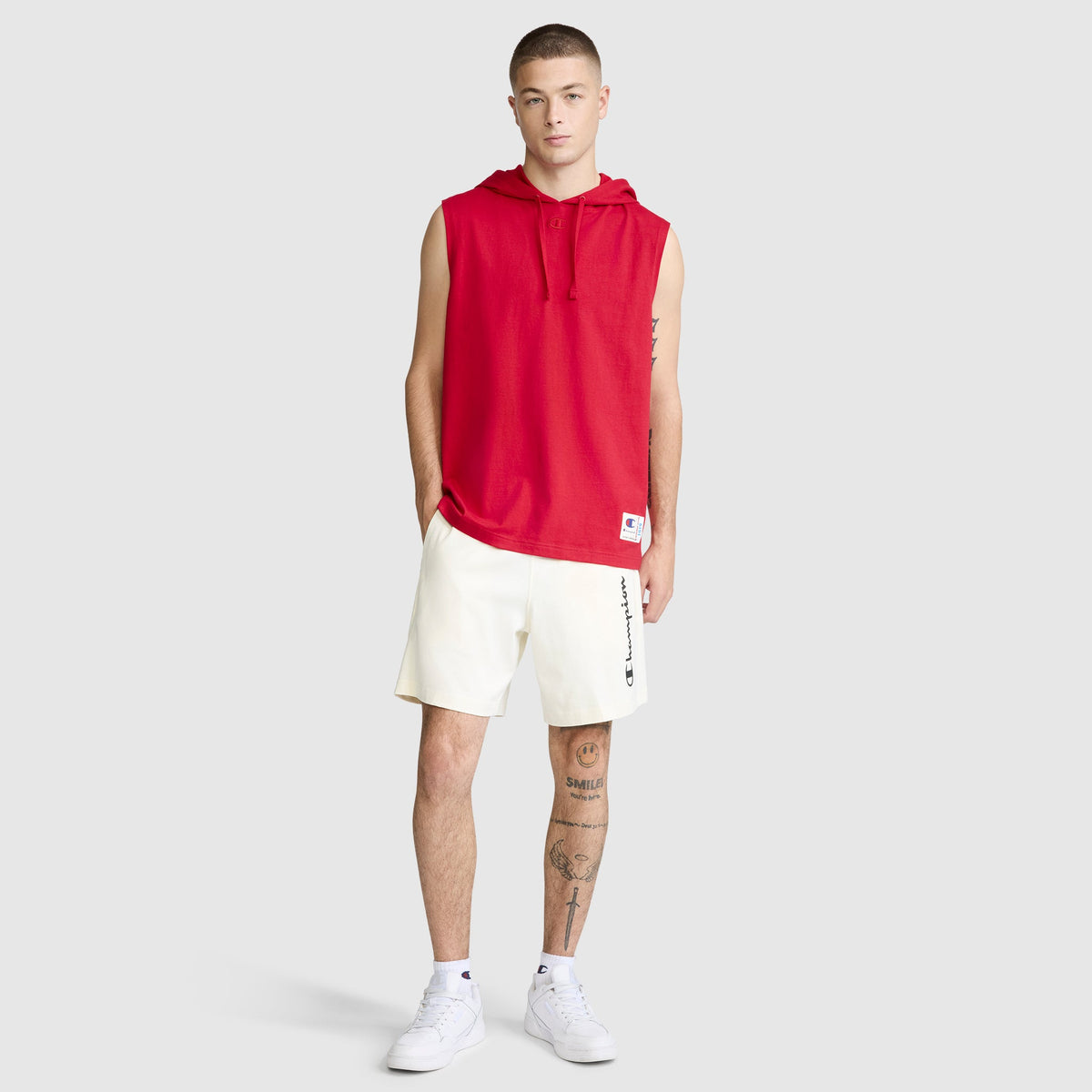 A young man stands against a plain light background, sporting a red Champion Jersey Sleeveless Hoodie with the C Logo and white shorts featuring the Champion text. He completes the look with white sneakers and displays tattoos on his left leg and right forearm.